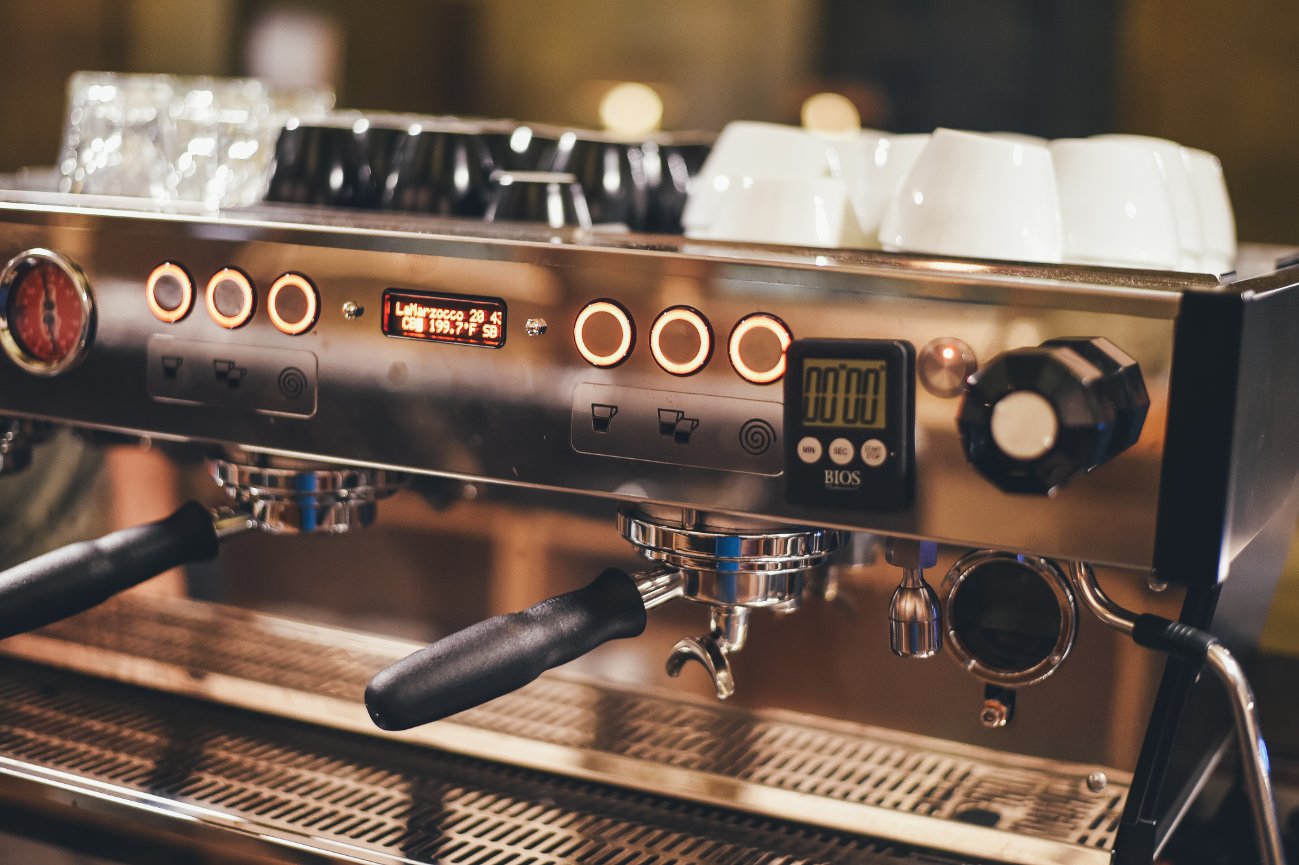 Espresso machine with digital displays and cup warmer, set in a warmly lit cafe environment.