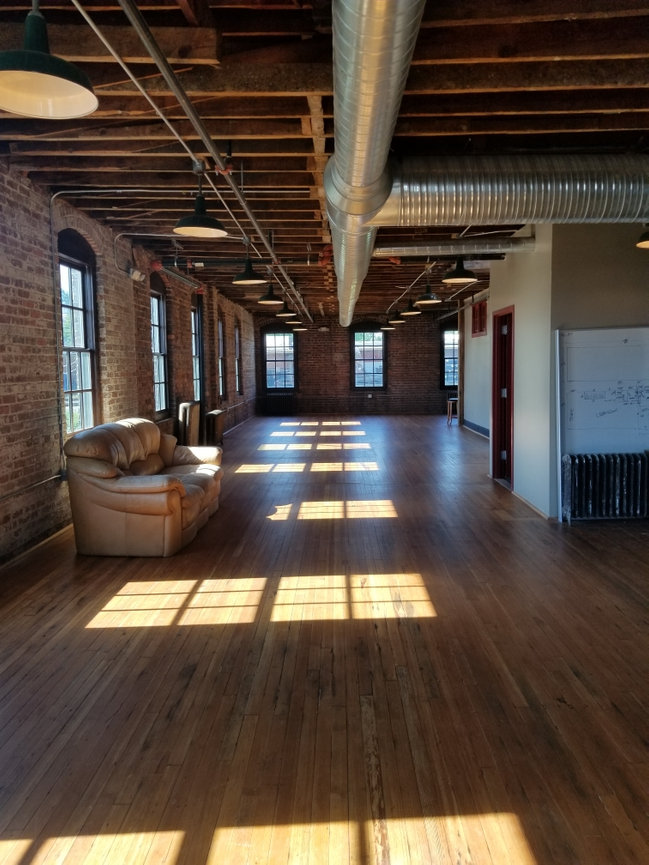 Loft with brick walls, wooden floors, leather couch, exposed beams, and ductwork. Bright sunlight streaming through windows.